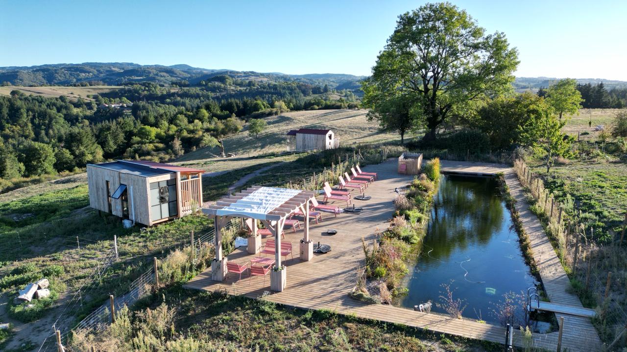 Le domaine vu du ciel : mini-maison en bois, pergola ombragée, terrasse et piscine naturelle au milieu des prés